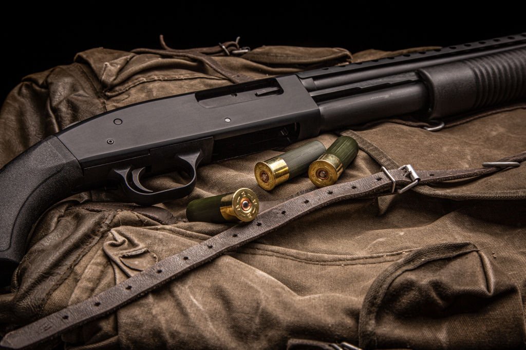 A black 12-gauge pump-action shotgun and ammunition resting on a canvas backpack. The shotgun features a plastic stock and is a smooth-bore weapon. The image has a dark background.